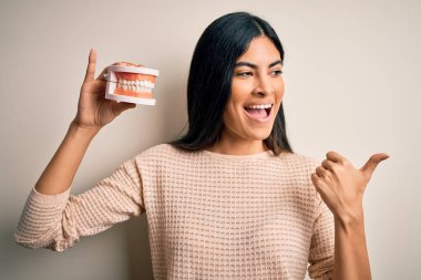 Diş protezi tutan genç ve güzel bir İspanyol dişçi. Ağzı protez ve baş parmağıyla işaret ediyor ve gülümsüyor.