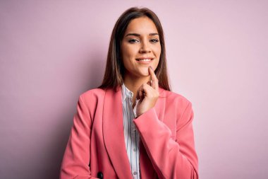 Pembe arka planda duran ceketli genç, güzel esmer kadın kameraya kollarını kavuşturup gülümseyerek ve çenesini kaldırarak bakıyor. Olumlu düşünüyorum..