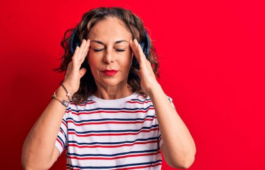 Orta yaşlı esmer kadın müzik dinliyor. Kırmızı arka planda kulaklık kullanıyor. Başı ağrıyor çünkü stres altında. Migren ağrısı..