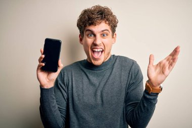 Sarışın, kıvırcık saçlı, akıllı telefonlu genç adam izole edilmiş beyaz arka planda mutlu ve heyecanlı duruyor. Kazanan ifade zaferi büyük bir gülümsemeyle kutluyor ve ellerini kaldırıyor.