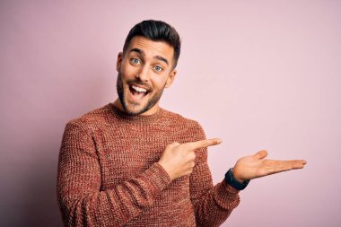 İzole edilmiş pembe arka plan üzerinde duran gündelik süveter giyen genç yakışıklı adam hayrete düşmüş ve kameraya gülümserken eliyle ve parmağıyla işaret ediyordu..