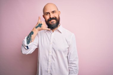Sakallı, zarif bir gömlek giyen, pembe arka planda gülümseyen, telefonla el ve parmaklarıyla konuşan, telefonda konuşan yakışıklı kel bir adam. İletişim kavramları.