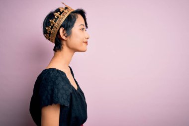 Genç, güzel Çinli bir kadın, kralın altın tacını takıyor, pembe arka planda, yan yana bakıyor, rahat profil pozu veriyor, kendine güvenen bir gülümsemeyle doğal bir poz veriyor..