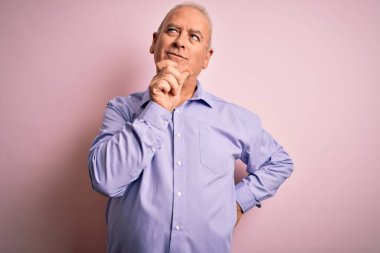 Orta yaşlı yakışıklı, beyaz gömlekli, pembe arka planda duran, elleri çenesinde soru soran, düşünceli bir adam. Düşünceli bir yüzle gülümsüyor. Şüphe kavramı.