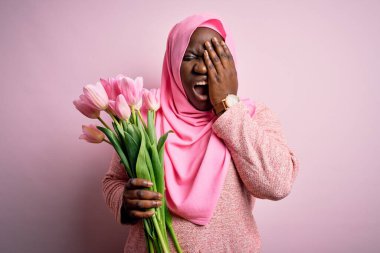 Genç Afro-Amerikan artı cüsseli kadın elinde pembe lale buketi tutan Müslüman başörtüsü tutan yarı yüzünü, gözünü ve ağzını eliyle kapatmaktan yorulmuş. Yüzüm acıyor..
