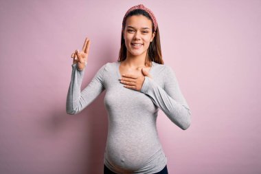 Genç, güzel bir kız hamile, pembe arka planda bebek bekliyor. Gülümsüyor, yemin ediyor, el pençe divan durarak yemin ediyor.