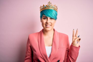 Mavi saçlı, pembe izole edilmiş arka planda kraliçe tacı takan genç bir iş kadını. Kameraya bakıp gülümsüyor ve zafer işareti yapıyor. İki numara..