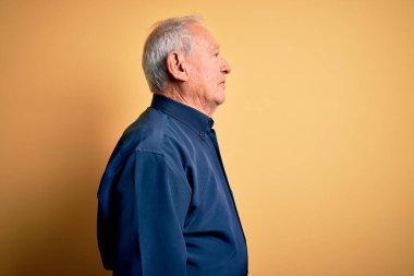 Gri saçlı, mavi gömlekli, sarı arka planda dikilmiş, rahat bir görünüm takınan, kendine güvenen bir gülümseme takınan yaşlı bir adam..