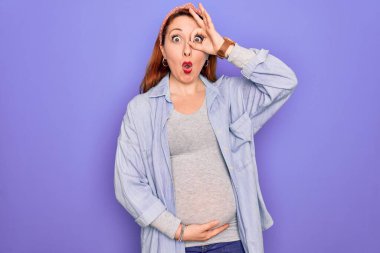 Genç, güzel, kızıl saçlı hamile bir kadın mor arka planda bebek bekliyor. Şok olmuş bir jest yapıyor. Şaşırmış bir yüz ve parmaklarından bakan bir gözle bakıyor. İnançsız ifade.
