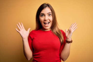 Sarı arka plan üzerine gündelik kırmızı tişört giyen güzel genç bir kadın çılgın ve başarıyı kollarını kaldırıp heyecanla haykırarak kutluyor. Kazanan konsept