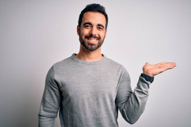 Sakallı, sıradan bir süveter giyen, beyaz arka planda durup gülümseyen, eli avucunda kameraya bakan genç ve yakışıklı bir adam..