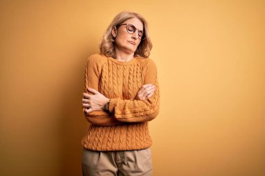 Orta yaşlı güzel sarışın kadın sarı arka planda günlük süveter ve gözlük takıyor. Kış soğuğu yüzünden titriyor ve donuyor. Yüzünde üzgün ve şok ifadesi var.