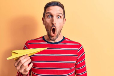 Sarı kâğıt uçak tutan yakışıklı genç adam izole arka planda duruyor ve ağzı açık, korkmuş ve şaşkın bir şekilde şaşkınlık içinde, inançsız bir yüz.