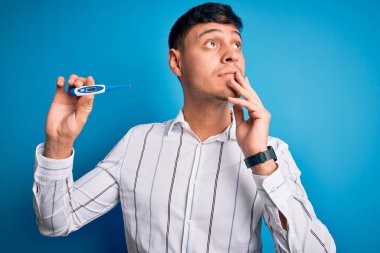 Tıbbi termometreyi mavi izole arka planda tutan genç İspanyol adam soru soran ciddi bir yüz, çok karışık bir fikir.