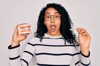 Kıvırcık saçlı ve gözlük takan güzel Afro-Amerikan kadın. Plastik takma dişler ve izole edilmiş beyaz arka plan üzerinde görünmez hizalayıcı tutuyor.