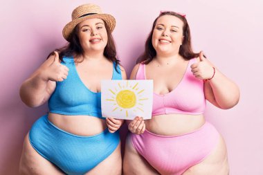 Bikini giyen genç ve büyük bedenli ikizler gülümseyen ve pozitif gülümseyen, başparmağıyla mükemmel ve onay işareti yapan insanlar. 