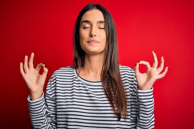 Genç, güzel, esmer bir kadın, kırmızı arka planda günlük çizgili tişört giyiyor. Gözleri kapalı, meditasyon yaparken parmaklarıyla gülümsüyor. Yoga konsepti.