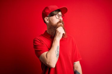 Gözlüklü yakışıklı teslimatçı ve izole edilmiş arka planda kırmızı şapkalı, elleri çenesinde soru sorarken, düşünceli bir yüz ifadesi vardı. Düşünceli bir yüzle gülümsüyor. Şüphe kavramı.