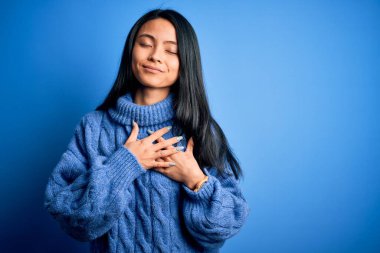 İzole edilmiş mavi arka plan üzerine günlük süveter giyen genç, güzel Çinli bir kadın elleri göğsünde, gözleri kapalı ve yüzünde minnet dolu bir jest ile gülümsüyor. Sağlık kavramı.