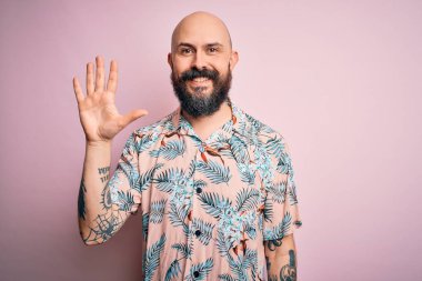 Sakallı ve dövmeli yakışıklı, pembe arka planda sıradan bir çiçek gömleği giyen ve gülümseyen, kendinden emin ve mutlu beş numaralı parmaklarıyla işaret eden kel adam..