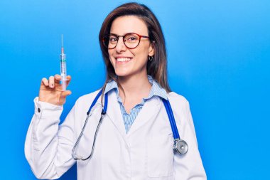 Doktor steteskobu takan genç ve güzel bir kadın elinde şırınga tutarken pozitif ve mutlu görünüyor. Kendine güvenen bir gülümsemeyle dişlerini gösteriyor. 