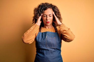 Orta yaşlı, güzel fırıncı kadın izole edilmiş sarı arka planda dikilmiş acı ve migrenden dolayı baş ağrısı ve stresten muzdarip önlük giyiyor. Ellerini başının üstüne koy..