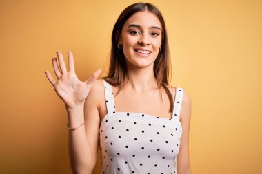 Sarı arka planda dikilen sıradan bir elbise giyen genç ve güzel esmer kadın beş numarayı gösterirken kendinden emin ve mutlu bir şekilde gülümsüyor..