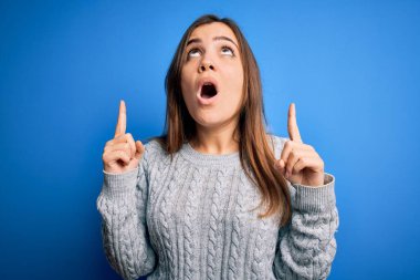 Mavi izole edilmiş arka plan üzerinde duran sıradan yün süveter giyen güzel genç bir kadın şaşırmış ve şaşırmış bir şekilde yukarı bakıyor ve parmaklarıyla işaret ediyor ve kollarını kaldırıyordu..