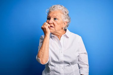 Soyutlanmış mavi arka plan üzerinde duran zarif tişört giyen son sınıf güzeli kadın elleri ağız yiyen tırnaklarla gergin ve stresli görünüyor. Endişe sorunu.