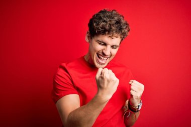 Sarışın, kıvırcık saçlı, kırmızı arka plan üzerine gündelik tişört giyen genç ve yakışıklı bir adam kolları havada ve gözleri kapalı olarak başarıyı kutluyor ve şaşırıyor. Kazanan konsept.