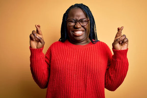 African american plus size woman with braids wearing casual sweater over yellow background gesturing finger crossed smiling with hope and eyes closed. Luck and superstitious concept.