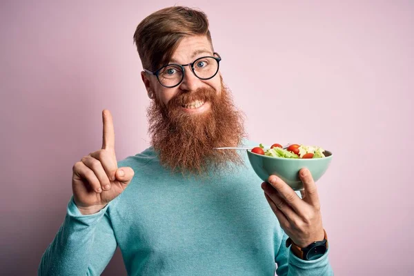 Redhead Irish healthy man with beard eating vegetarian green salad over pink background surprised with an idea or question pointing finger with happy face, number one