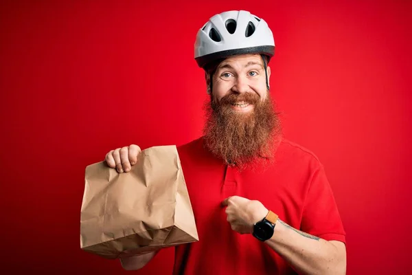 Redhead Irish delivery man with beard wearing bike helmet and holding takeaway paper bag with surprise face pointing finger to himself