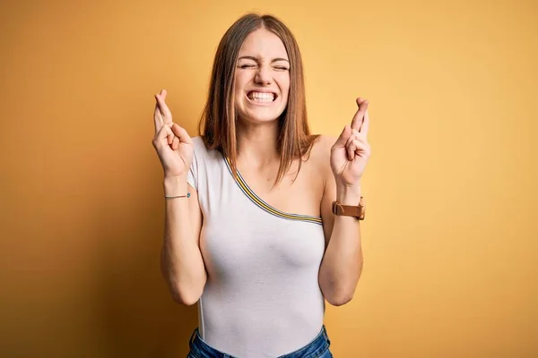 孤立した黄色の背景にカジュアルなTシャツを着た若い美しい赤毛の女性は 希望と目を閉じて笑顔を交差ジェスチャー指 幸運と迷信の概念 — ストック写真