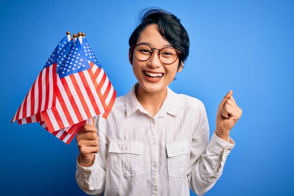 Young beautiful asian patriotic girl holding united states flags celebrating independence day screaming proud and celebrating victory and success very excited, cheering emotion