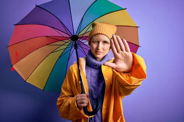 金发碧眼的年轻女子 身穿防水雨衣 头戴五颜六色的雨伞 张开双手 严肃而自信地做着止步手势 做着防守动作 — 图库照片