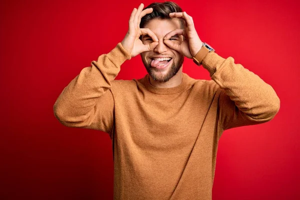 Sakallı ve mavi gözlü genç bir adam kırmızı arka planda sıradan bir süveter giyiyor. Dürbün gibi jest yapıyor. Dürbün dilini çıkarıyor, gözler parmaklarından bakıyor. Çılgın ifade.