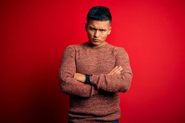 Young handsome latin man wearing casual sweater standing over red background skeptic and nervous, disapproving expression on face with crossed arms. Negative person.