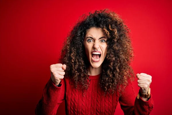 Young beautiful woman with curly hair and piercing wearing casual red sweater angry and mad raising fists frustrated and furious while shouting with anger. Rage and aggressive concept.