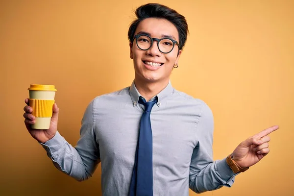 Young handsome chinese man drinking glass of take away coffee over yellow background very happy pointing with hand and finger to the side