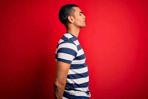英俊的非洲裔美国男子 身穿休闲条纹T恤 站在红色背景上 向侧面看去 以自然的面容和自信的微笑放松自己的姿态 — 图库照片