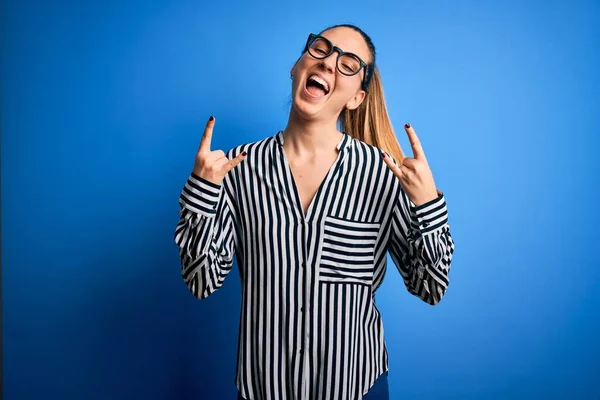 Beautiful blonde woman with blue eyes wearing striped shirt and glasses over blue background shouting with crazy expression doing rock symbol with hands up. Music star. Heavy concept.