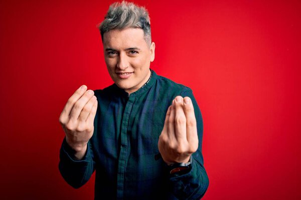 Young handsome modern man wearing elegant green shirt over red isolated background doing money gesture with hands, asking for salary payment, millionaire business