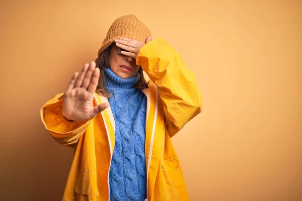 Middle age woman wearing yellow raincoat and winter hat over isolated background covering eyes with hands and doing stop gesture with sad and fear expression. Embarrassed and negative concept.