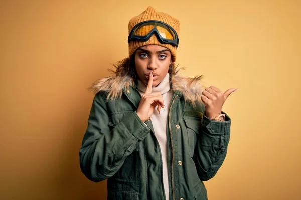 身穿雪地运动服和滑雪护目镜的年轻的非洲裔美国滑雪者女孩要求保持安静 手指放在嘴唇上 手拉手指向侧边 沉默和秘密概念 — 图库照片
