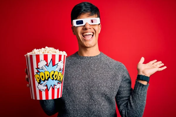 Young handsome man watching movie eating popcorns snack over isolated red background very happy and excited, winner expression celebrating victory screaming with big smile and raised hands