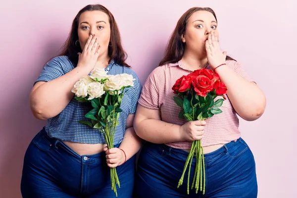 Young plus size twins holding flowers covering mouth with hand, shocked and afraid for mistake. surprised expression