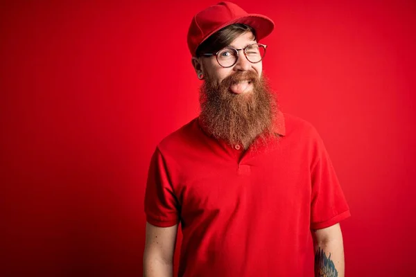 Young handsome delivery man wearing glasses and red cap over isolated background winking looking at the camera with sexy expression, cheerful and happy face.