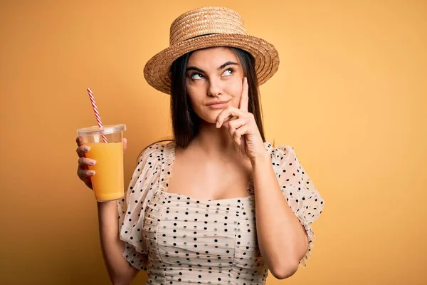 年轻美丽的黑发女子戴着夏帽 喝着橙汁 脸上严肃地思考着问题 非常迷惑的想法 — 图库照片