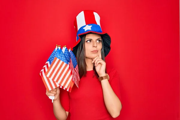 Young beautiful brunette patriotic woman wearing united states hat holding usa flags serious face thinking about question with hand on chin, thoughtful about confusing idea